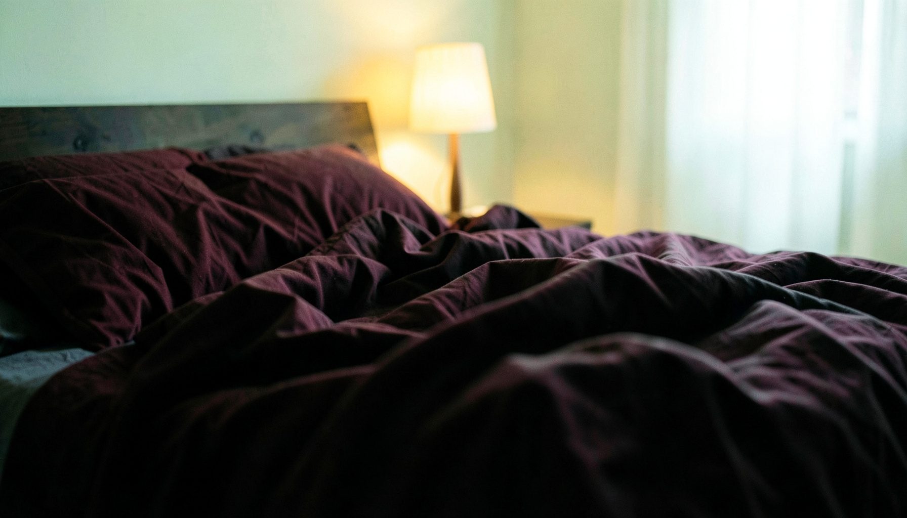 A dimly lit bedroom interior with rumpled bedding in deep burgundy and charcoal tones, warm amber light spilling from a bedside lamp, moody and intimate atmosphere